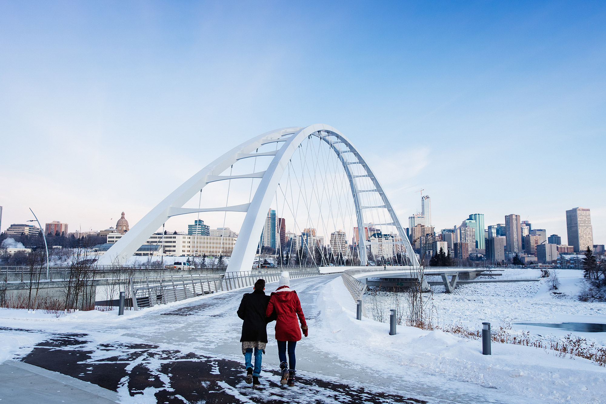 Je ziet twee vrouwen die bij de grote brug van Edmonton lopen. Overal ligt sneeuw en de lucht is helderblauw.